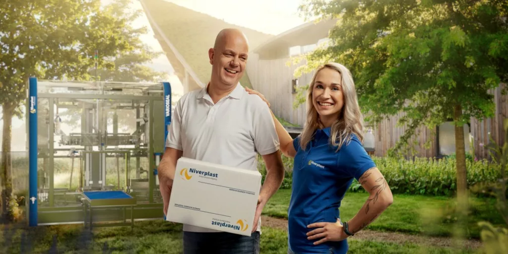 Twee medewerkers van Niverplast bij een verpakkingsmachine, één met een doos in de hand op het bedrijfsterrein.